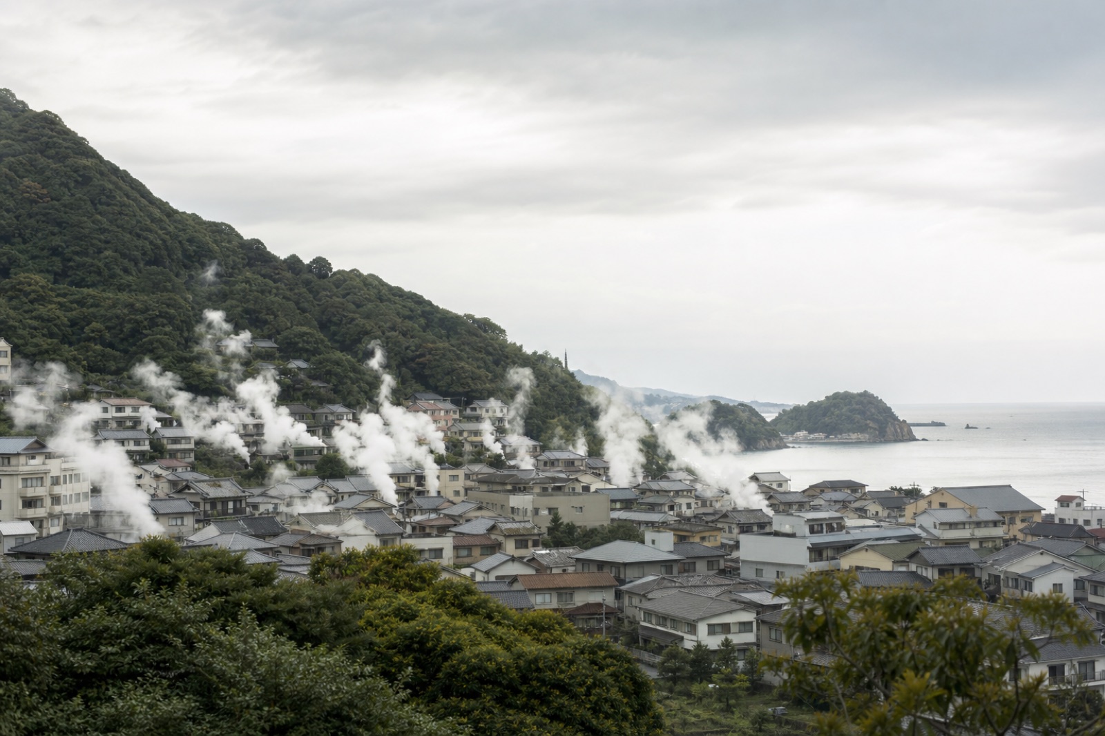 Ånga som stiger från många punkter över en japansk stad vid foten av kullar med havet bortom