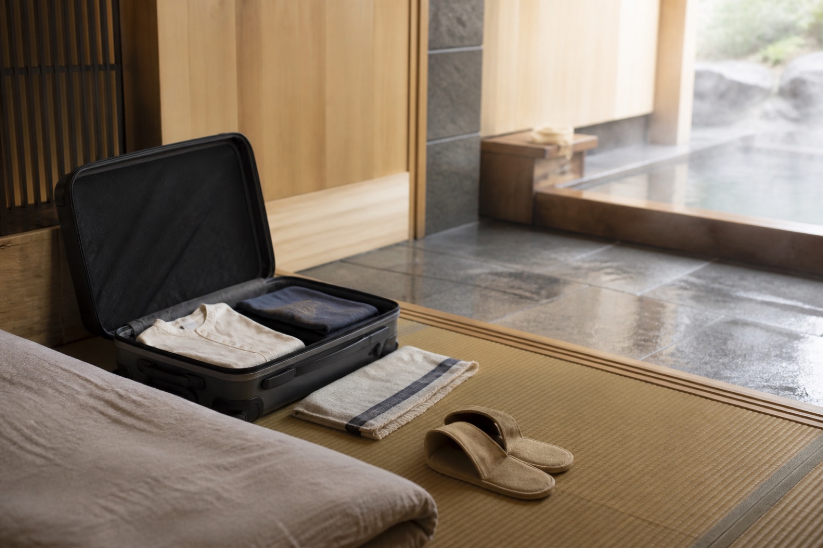 Öppen liten resväska med vikta kläder, en tunn handduk och tofflor på tatami